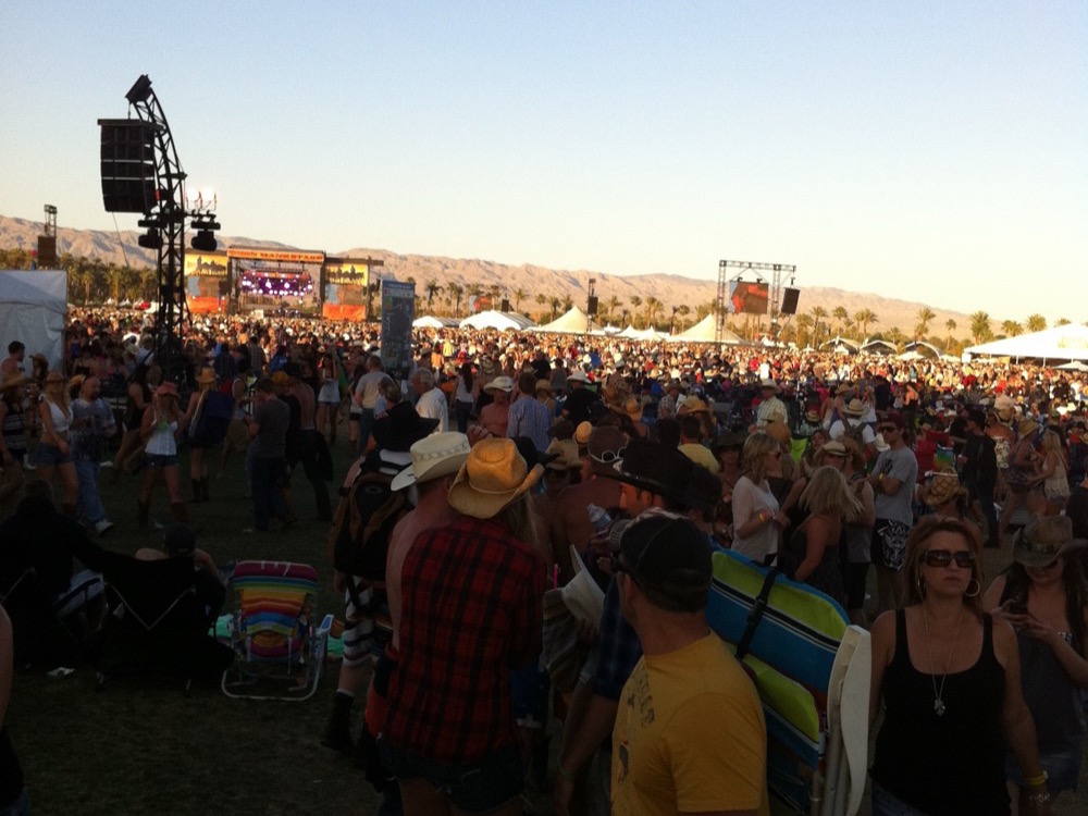 A crowd in cowboy hats at the Stagecoach festival in Indio at sunset, stages and palm trees behind