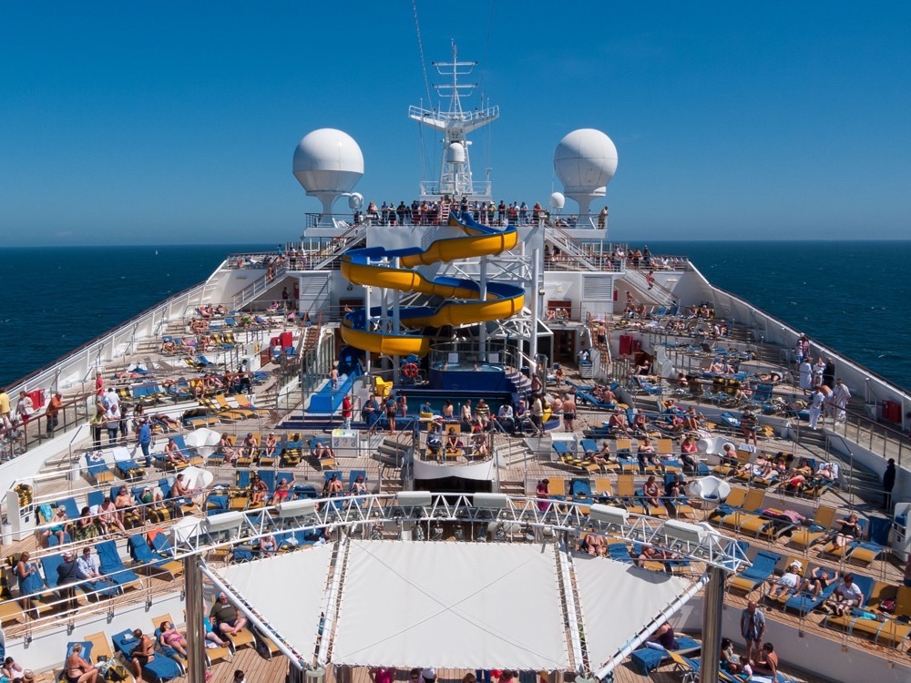 The open pool deck of a cruise ship at sea, with satellite domes above the bridge