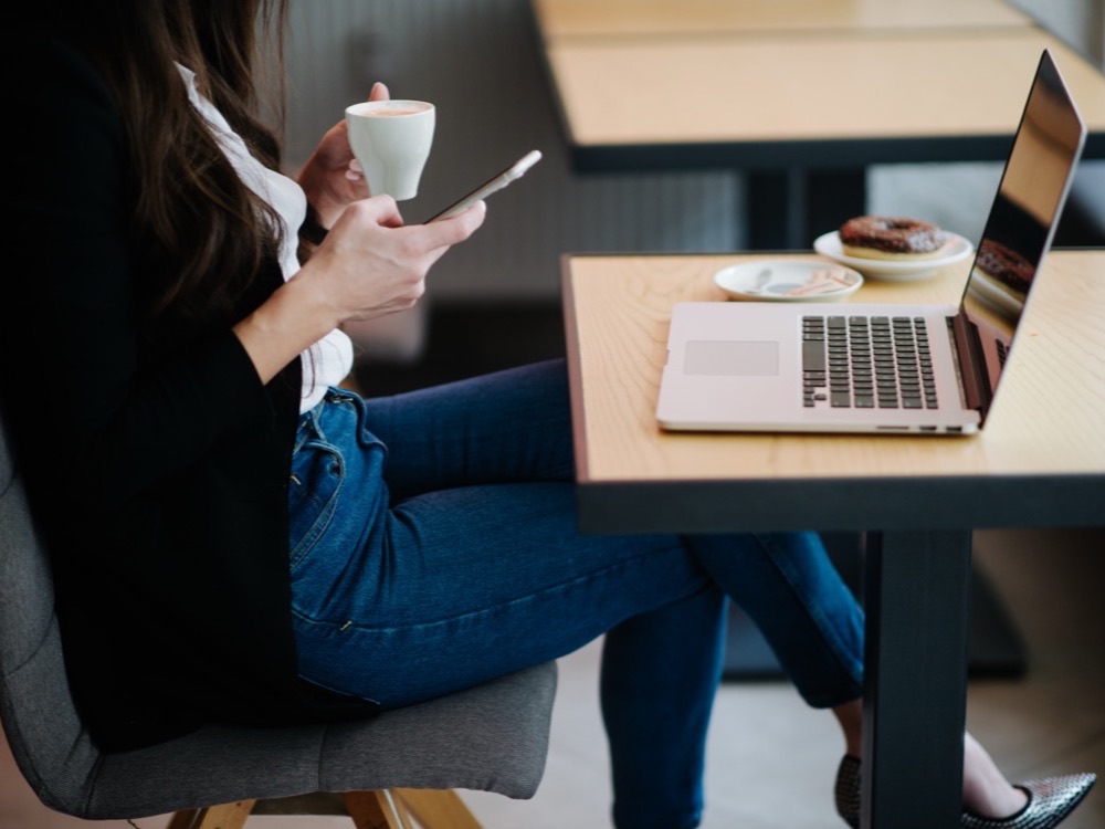 A woman in a café holding a coffee and a smartphone, a laptop open on the table