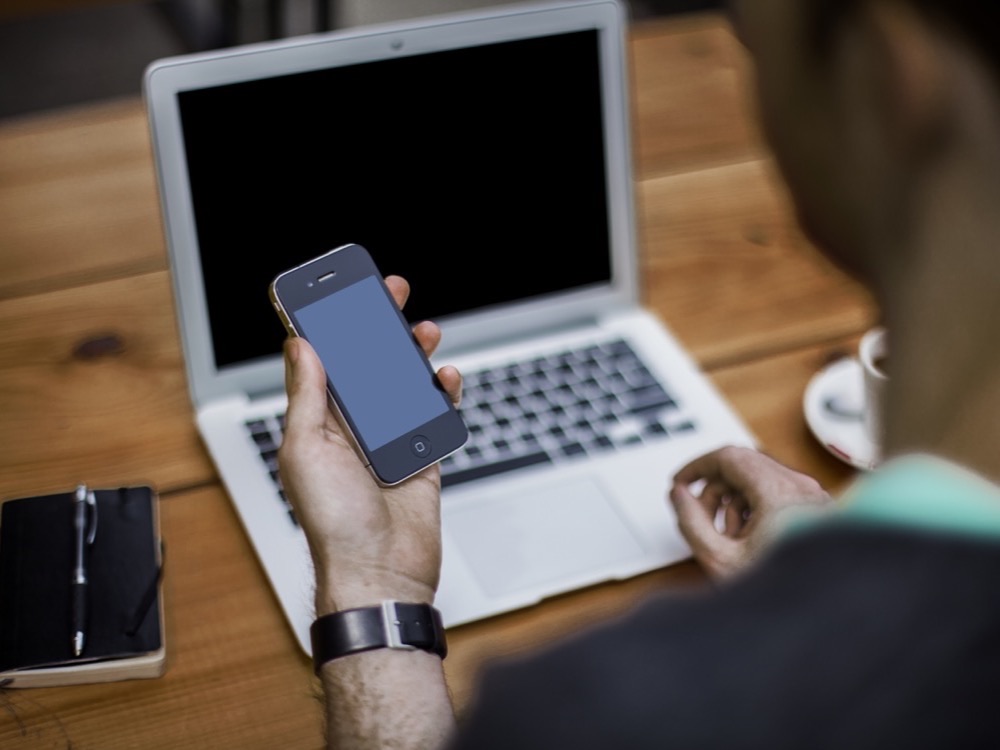 A man at a laptop holding a smartphone