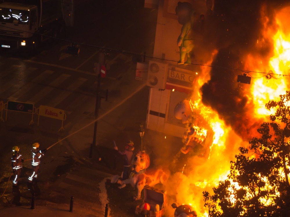 A falla monument burning at night in a Valencia street, firefighters standing by