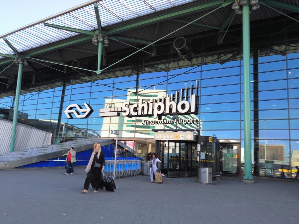 The glass entrance of Amsterdam Schiphol Airport under a blue sky, with the Schiphol sign above the doors